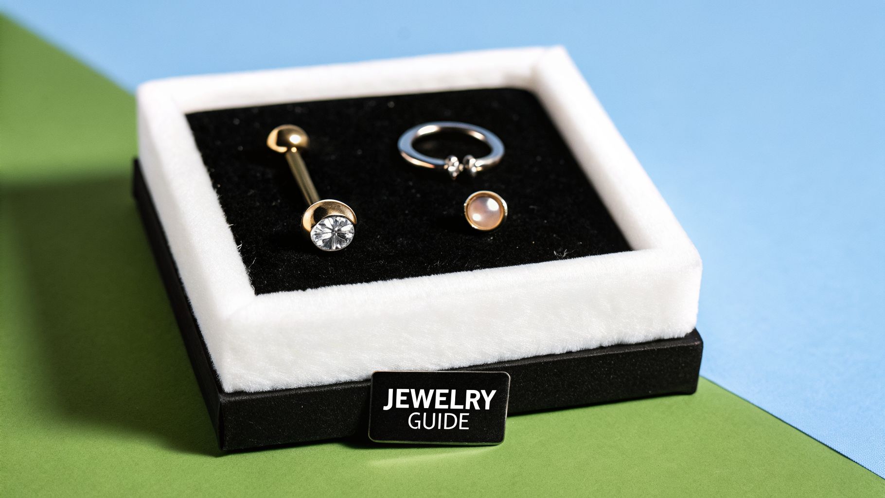 Black jewelry box with gold barbell piercing, silver ring, and pink stud on black velvet.