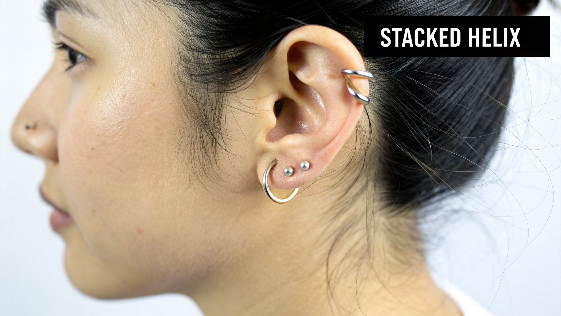 Close-up of a person's ear featuring a stacked helix, stud, and hoop piercings, plus a nose piercing.