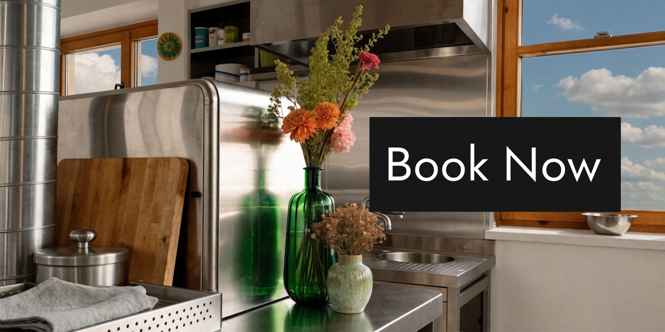 A stainless steel kitchen counter featuring a green glass vase with fresh flowers and a small pot.