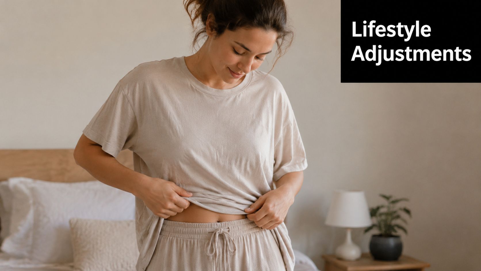 A woman wearing beige loungewear looking down at her stomach in a softly lit bedroom.