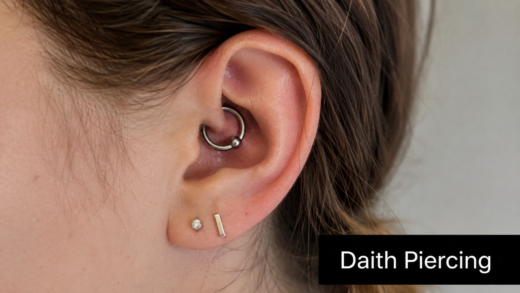 Close-up of a human ear showing a metal hoop daith piercing and two small lobe earrings.