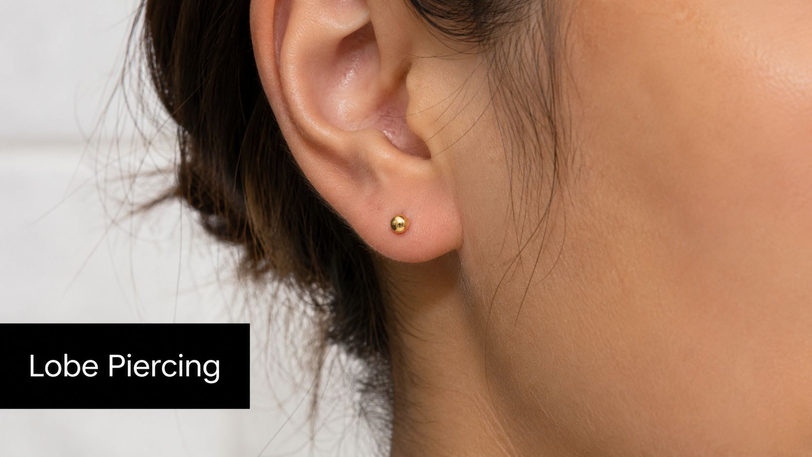 A close-up view of a woman's ear featuring a simple and elegant gold ball lobe piercing.