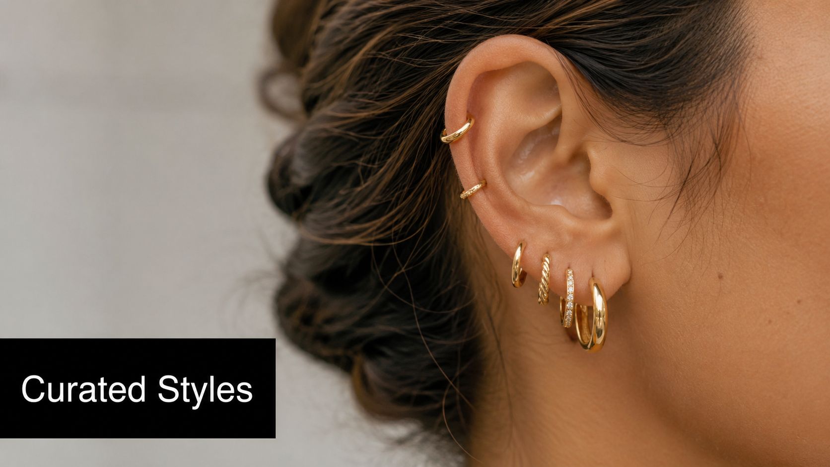 A close-up of a woman's ear wearing a stack of multiple delicate small gold hoop earrings.