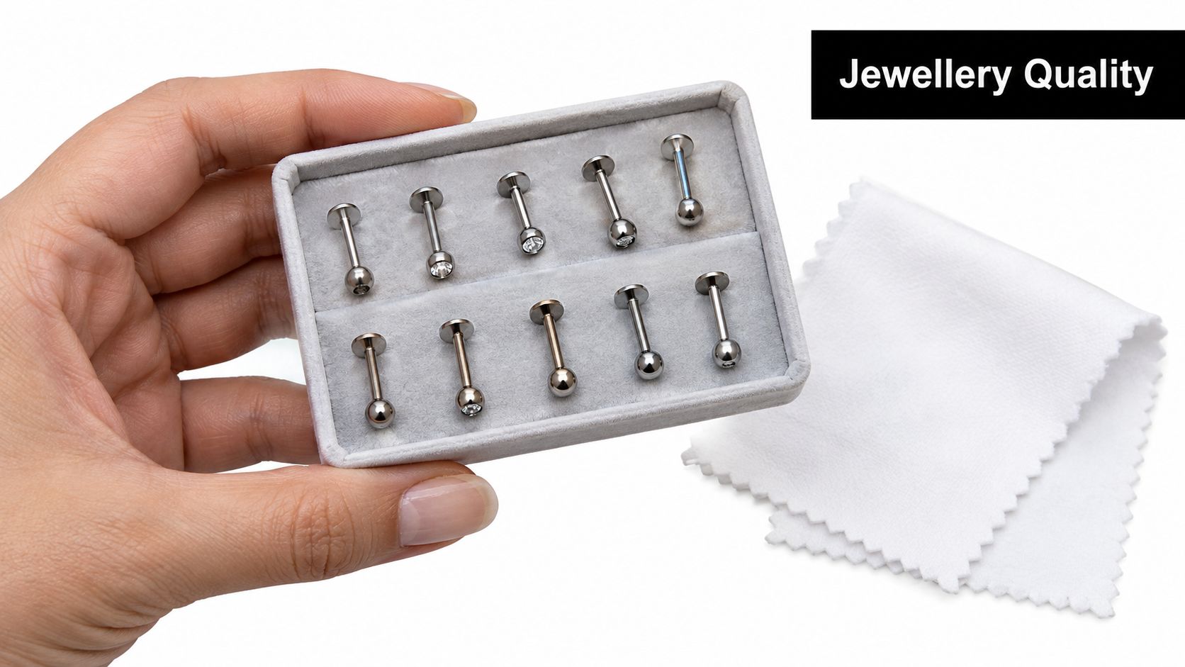 A hand holding a small grey display box filled with ten silver labret piercing studs and a cloth.